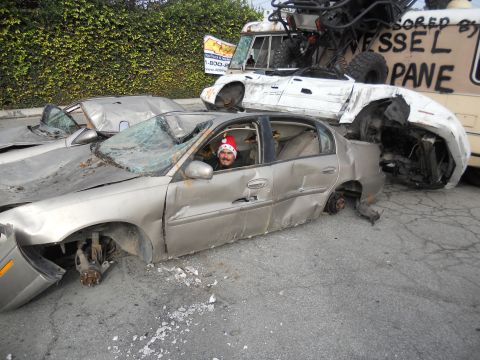 1986 Ford Bronco - A man in a Santa hat sits in a heavily damaged silver sedan, surrounded by other wrecked vehicles in a junkyard.
