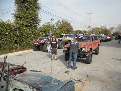 1986 Ford Bronco - An orange pickup truck is parked in a line with other off-road vehicles, with a person working on its front end.
