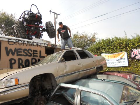 1986 Ford Bronco - Junkyard scene shows a buggy on a trailer, a man standing on a sedan, and other cars piled up.