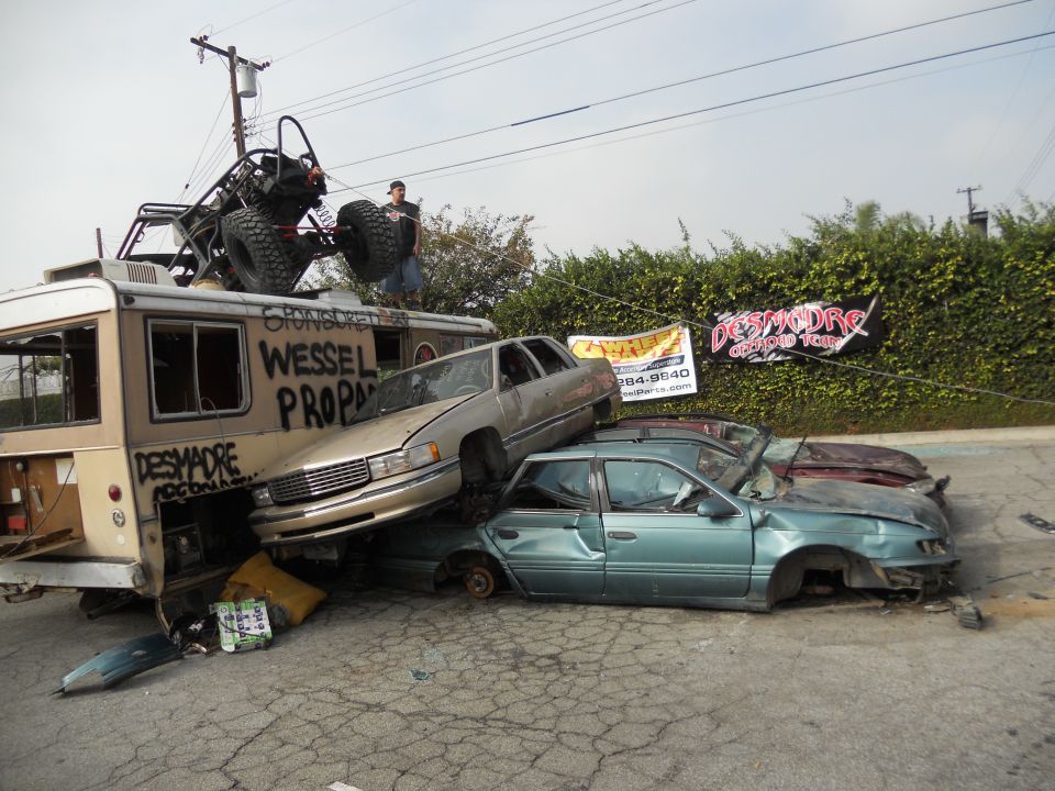 Bus with buggy on roof, gold sedan on top of teal sedan, red car behind teal sedan. Banners and graffiti on bus