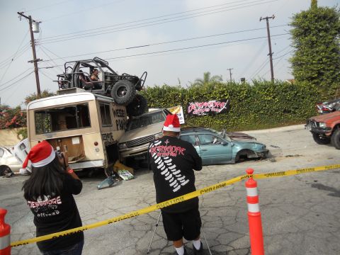 1986 Ford Bronco - A bizarre scene unfolds with multiple damaged vehicles, a bus carrying a buggy, and individuals in festive attire.