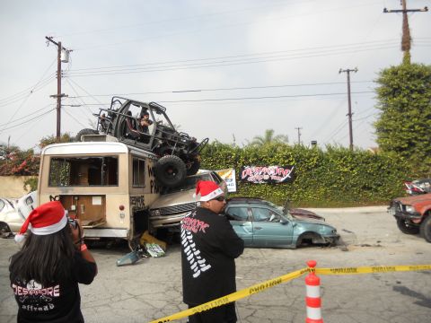 1986 Ford Bronco - A chaotic scene with a buggy stacked on a bus, surrounded by damaged vehicles and onlookers in festive hats.