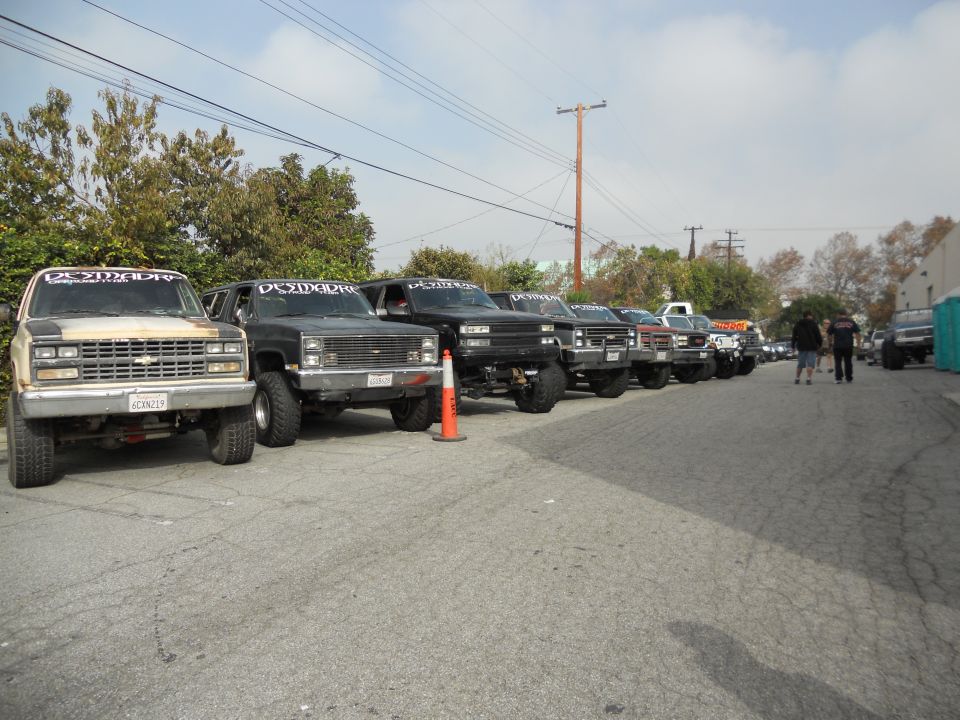 Several lifted pickup trucks and SUVs parked in a line. The first truck has a 'DESMADRE' banner on its windshield