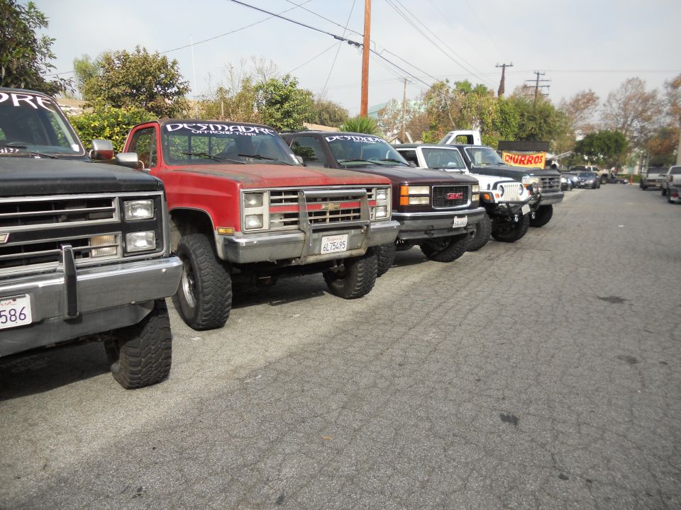 Several lifted trucks parked in a row, front view. The red truck has a "DESMADRE OFFROAD TEAM" banner