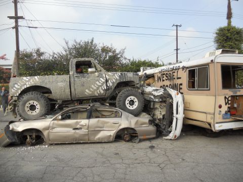 1986 Ford Bronco - A heavily modified pickup truck has crushed a sedan, with a bus visible in the background. Text on the bus indicates sponsorship by 'WESSEL…