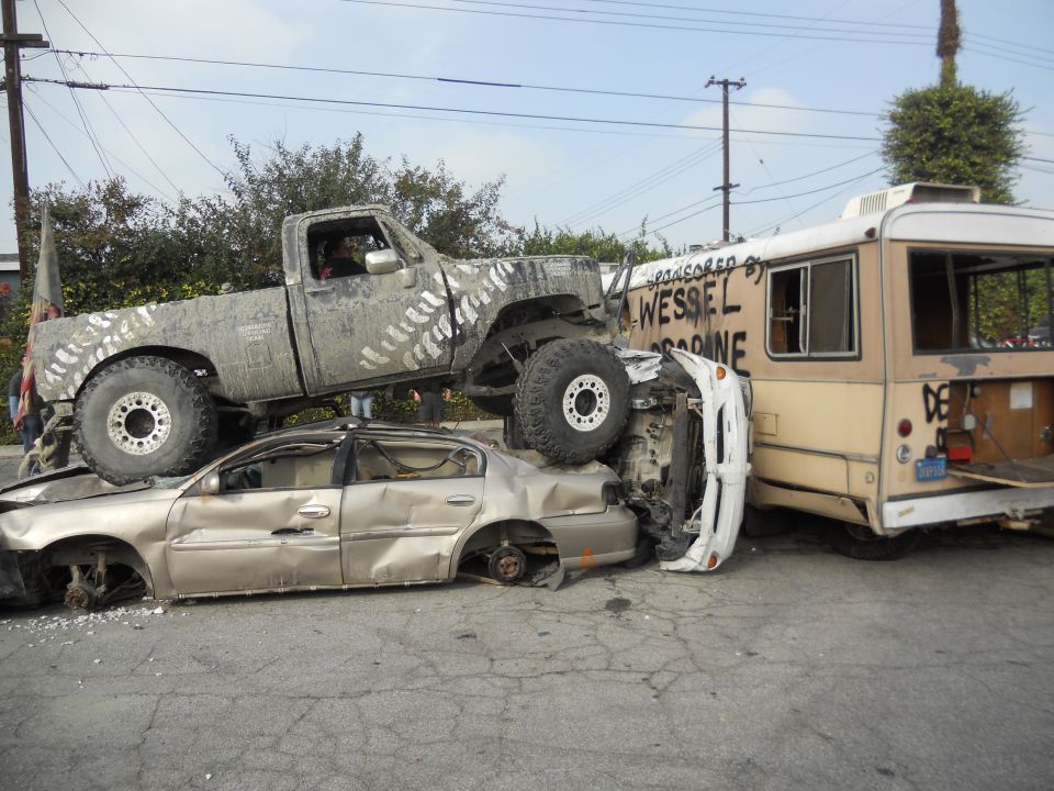 Truck on top of a sedan, next to a bus. All look wrecked