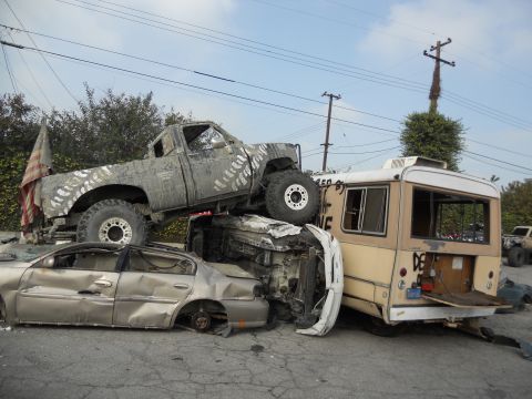 1986 Ford Bronco - A chaotic scene shows a heavily modified pickup truck perched atop other vehicles, with an American flag attached to its rear.