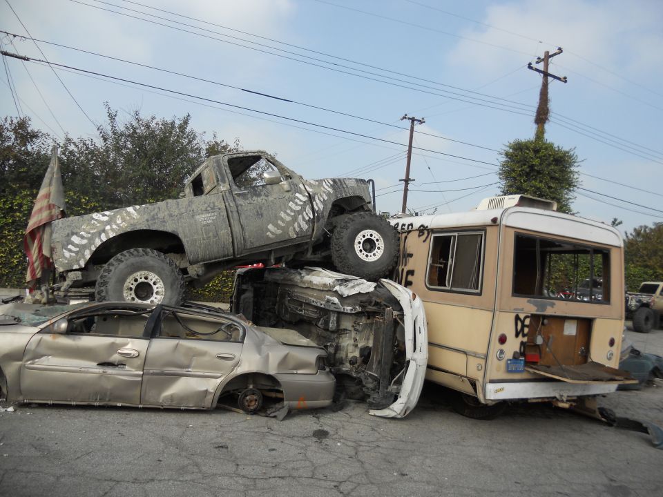 Mud-splattered pickup truck on top of a crushed sedan and a beige bus