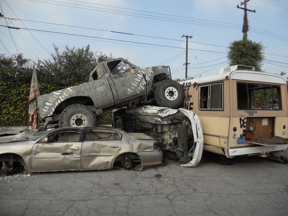 Wrecked pickup truck on top of sedan, next to a bus. Tire tracks painted on truck