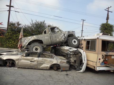 1986 Ford Bronco - A chaotic scene with multiple vehicles in various states of disrepair, including a lifted truck piled on top of other cars.