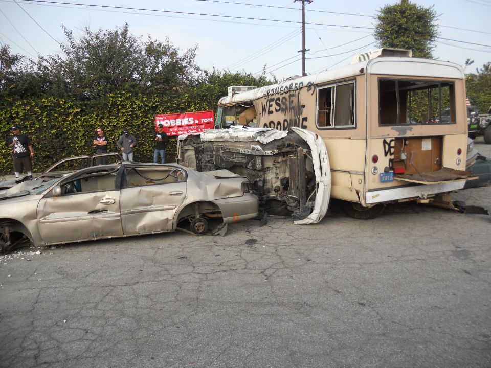 wrecked sedan and bus on street, people standing around, banner in background