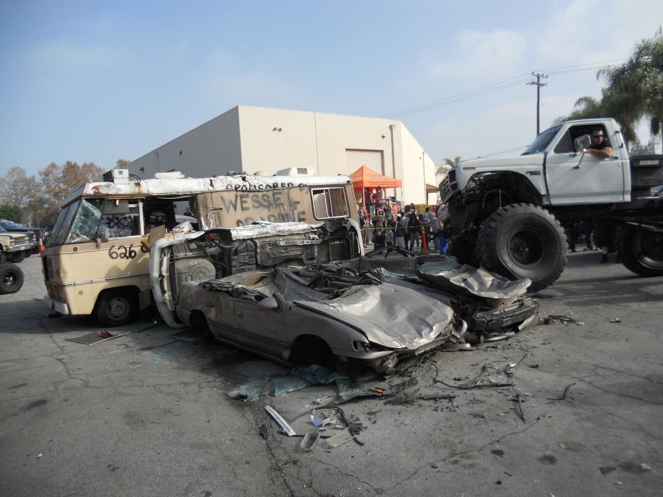 Wrecked van and car crushed by a monster truck. Text on van says 'WESSEL OF HOME'