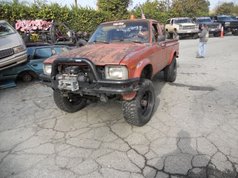 1986 Ford Bronco - A modified red pickup truck sits in a lot with other vehicles and a banner in the background.
