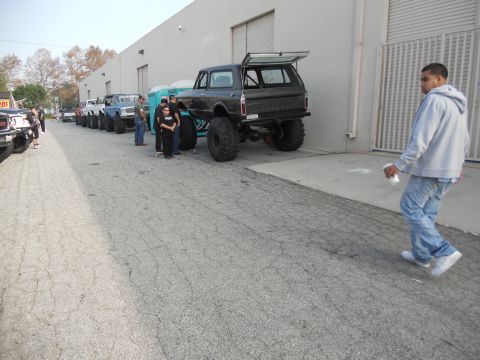 1986 Ford Bronco - A line of modified off-road vehicles is parked outside a commercial building.