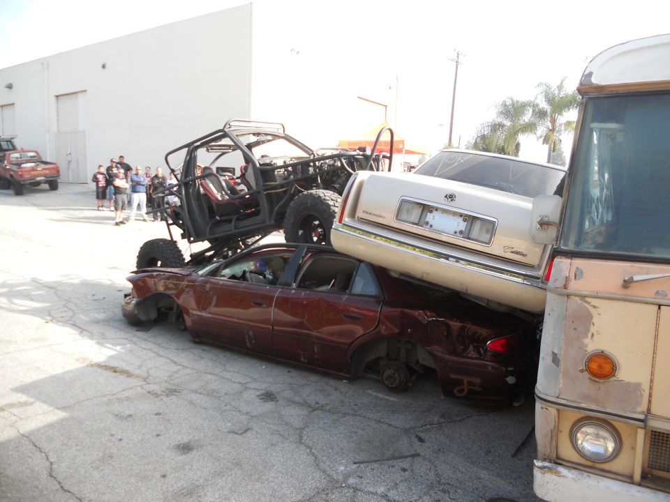Several wrecked cars are stacked, with a custom off-road vehicle on top. A bus is parked to the right