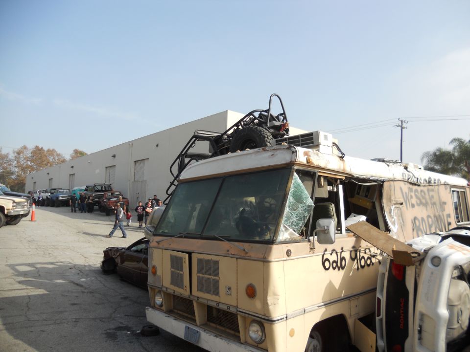 Front view of a tan custom RV with a roll cage and spare tire on the roof, parked in a lot with other vehicles
