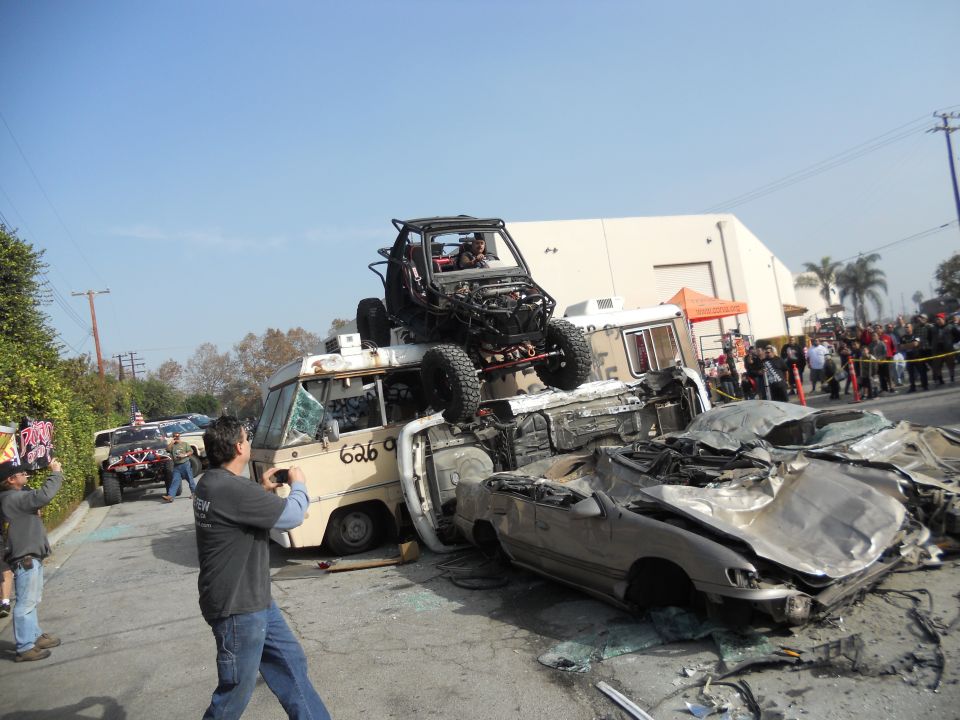 Off-road buggy atop a pile of wrecked cars and a van, with spectators watching