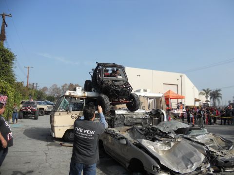 1986 Ford Bronco - An off-road buggy is precariously perched atop a stack of damaged vehicles, with onlookers gathered.
