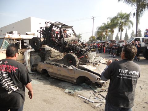 1986 Ford Bronco - An off-road buggy is precariously balanced on a pile of wrecked vehicles, drawing a crowd of onlookers.