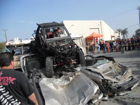 1986 Ford Bronco - An off-road buggy is positioned on top of a severely damaged car, with spectators gathered around.