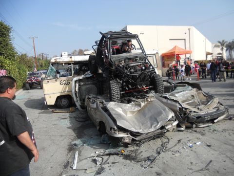 1986 Ford Bronco - A chaotic scene with an off-road buggy perched atop severely damaged cars, a van nearby, and onlookers observing.