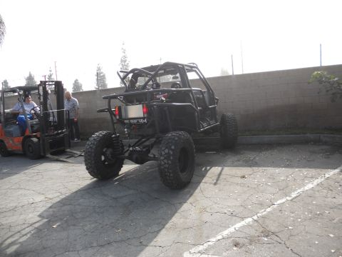 1986 Ford Bronco - A custom-built buggy with a prominent roll cage and oversized tires is parked outdoors next to a forklift.