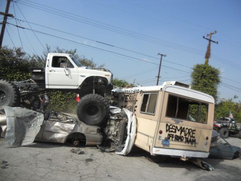1986 Ford Bronco - Modified pickup truck rests precariously atop a wrecked bus and car, with 'DESMADRE OFF ROAD TEAM' spray-painted on the bus.