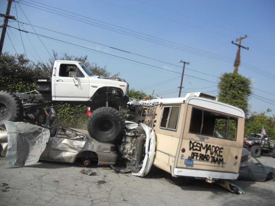 White pickup truck with large tires is on top of a wrecked bus and car. Text on bus reads 'DESMADRE OFF ROAD TEAM'
