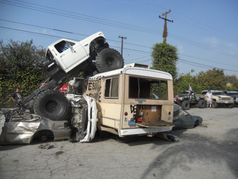 1986 Ford Bronco - A white pickup truck is precariously balanced on a bus, which is itself crushing several cars.