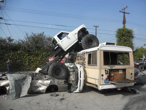 1986 Ford Bronco - A chaotic scene shows a modified pickup truck precariously perched atop a bus, with a car crushed beneath them.