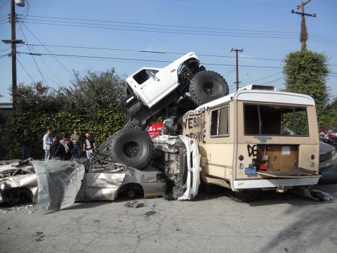 1986 Ford Bronco - A chaotic scene with a monster truck crushing a bus and car, surrounded by onlookers.