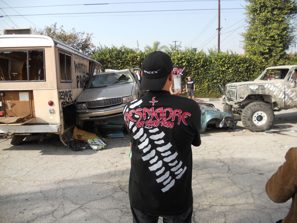 Person in a 'DESMADRE OFFROAD TEAM' shirt looks at a scene with a wrecked bus, a damaged sedan, and a large off-road truck