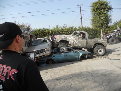 1986 Ford Bronco - A heavily modified pickup truck is parked on top of two wrecked cars, with a person filming the scene.