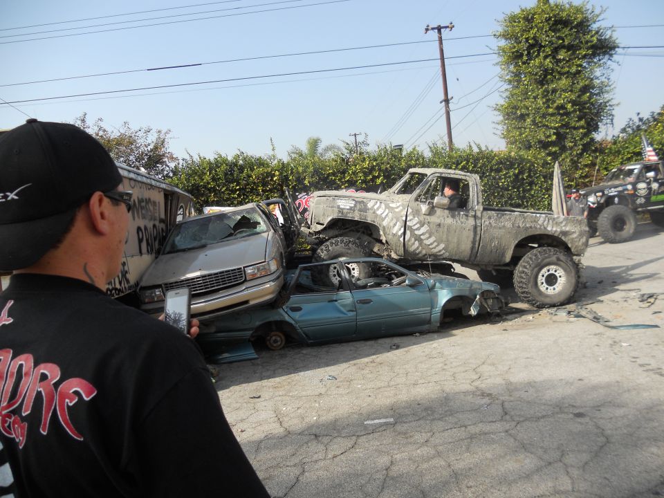 Mud-splattered pickup truck on top of crushed cars, with a person filming