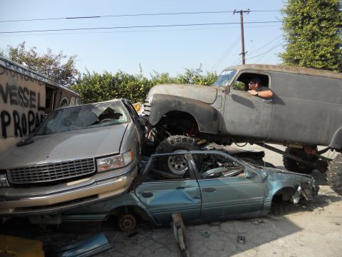 1986 Ford Bronco - A heavily modified panel van is piled on top of two heavily damaged sedans in a junkyard.