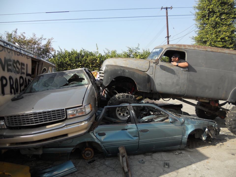 Gray panel van with large tires crushing a silver sedan and a blue sedan