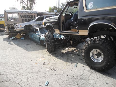 1986 Ford Bronco - A lifted 1986 Ford Bronco is parked on top of a wrecked sedan, with other vehicles in the background.