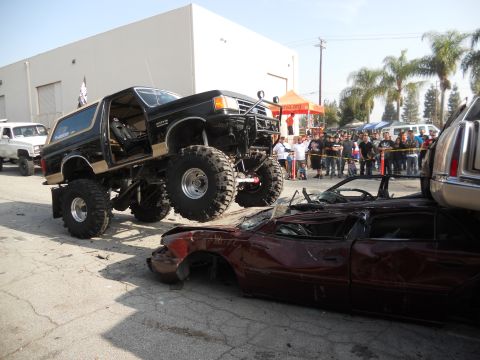 1986 Ford Bronco - A heavily modified black Ford Bronco is shown crushing a red car, with a crowd watching.