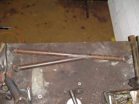 1993 Ford Bronco - Two long steel tube links with weld-on end bungs lie on a shop table, joined near the middle by a welded cross-brace to form a triangulated…