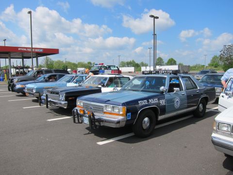 1991 Ford LTD Crown Victoria - dark blue Ford LTD Crown Victoria "State Police" sedan with push bar leads a row of classic cars in a parking lot under a cloudy sky.