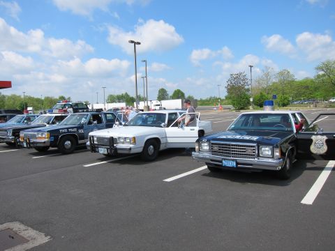 1991 Ford LTD Crown Victoria - lineup of vintage police vehicles, including State Police sedans and a pickup, parked diagonally in a parking lot under a partly cloudy sky.