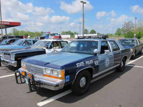 1991 Ford LTD Crown Victoria - two-tone blue Ford LTD Crown Victoria marked State Police 90E sits with a roof light bar at a sunny outdoor police-car gathering.