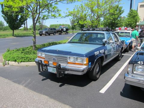 1991 Ford LTD Crown Victoria - faded blue State Police Ford Crown Victoria with front push bar and roof light bar parked among other cruisers on a sunny day.