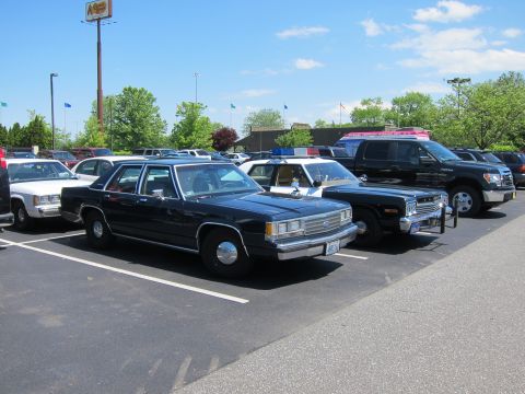 1991 Ford LTD Crown Victoria - lineup of vintage police cruisers, including dark Ford sedans and a black-and-white car, parked in a sunlit lot under a blue sky.