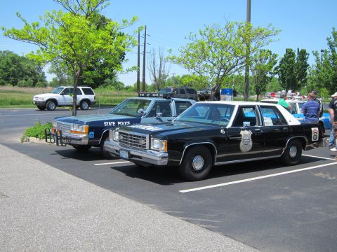 1991 Ford LTD Crown Victoria - black-and-white police Ford sedan marked 45 with a gold badge decal parked next to a blue State Police cruiser in a grassy lot.