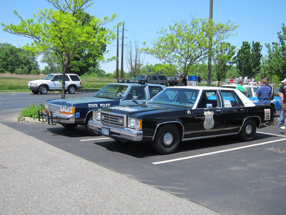 Black-and-white police Ford sedan number 45 with badge decal beside a blue State Police car