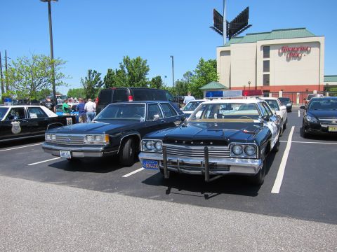 1991 Ford LTD Crown Victoria - black Dodge police sedan with chrome push bar sits beside a Ford cruiser in a hotel parking lot at a police-car gathering.
