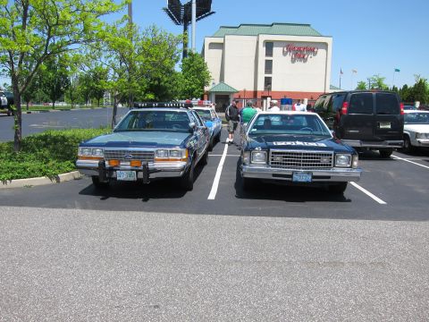 1991 Ford LTD Crown Victoria - blue Ford State Police cruiser and a black-and-white police sedan parked nose-out side by side near a hotel on a sunny day.