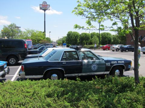1991 Ford LTD Crown Victoria - dark blue State Police 90E Ford cruiser with light bar parked behind a hedge, with a Dunkin Donuts sign in the background.