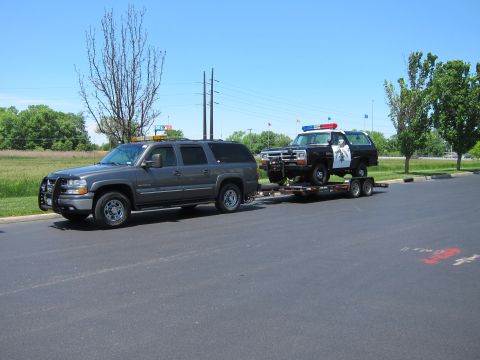 1991 Ford LTD Crown Victoria - gray Chevrolet Suburban tows a flatbed trailer carrying a black-and-white police SUV across an empty lot.